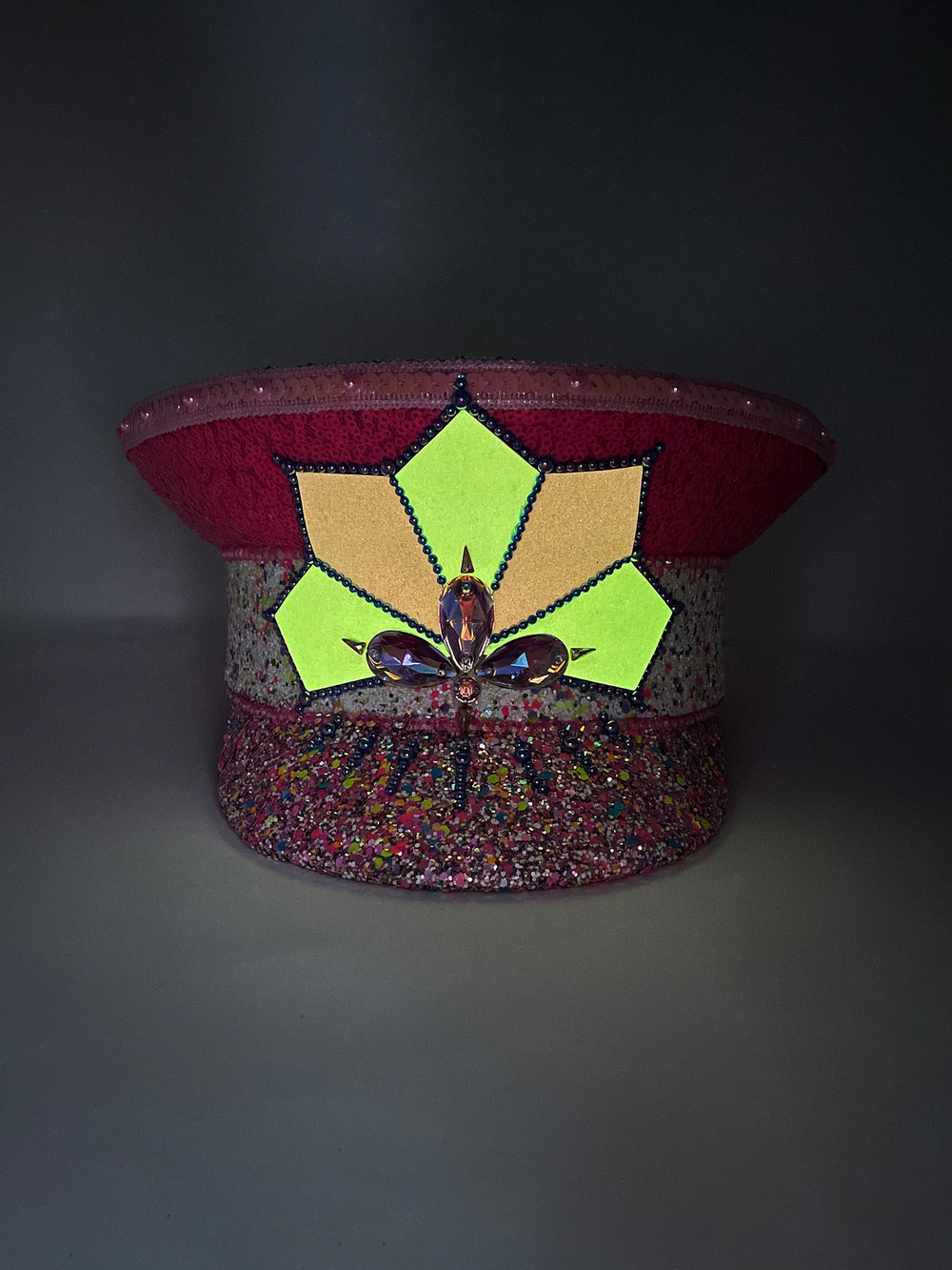 Festival captain hat with pink sequins, colourful glitter peak, Jukebox logo jewel detail, and glow-in-the-dark reflective starburst design – perfect for rave, costume, or party wear.
